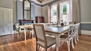 A dining room with a wooden table and chairs, a chandelier, and large windows with curtains. - Parker Cottage (Cape Town)