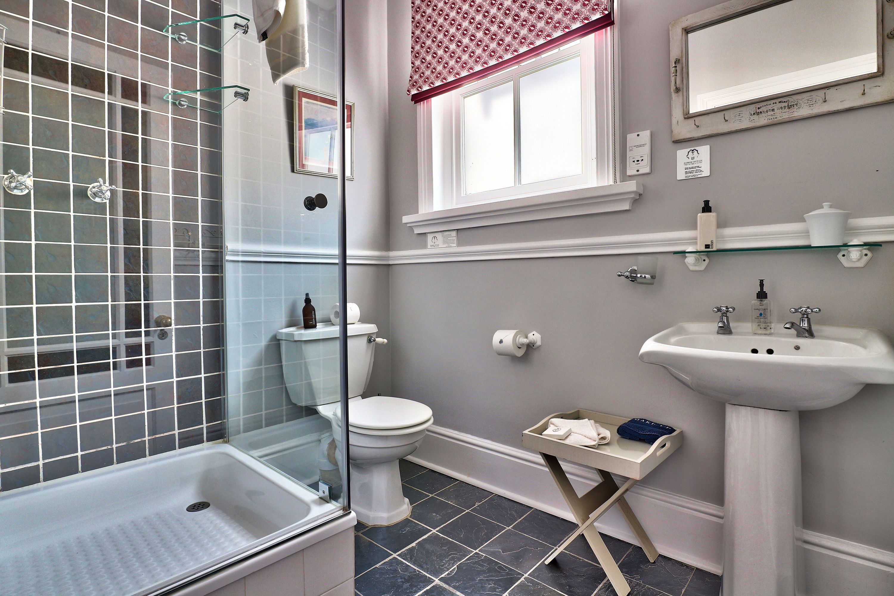 A bathroom with a glass shower enclosure, a toilet, a sink, and a window with a red valance.