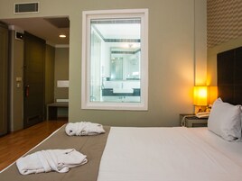 Ένα δωμάτιο ξενοδοχείου με ένα κρεβάτι, δύο πετσέτες, ένα κομοδίνο με ένα φωτιστικό και θέα στο μπάνιο από ένα παράθυρο.