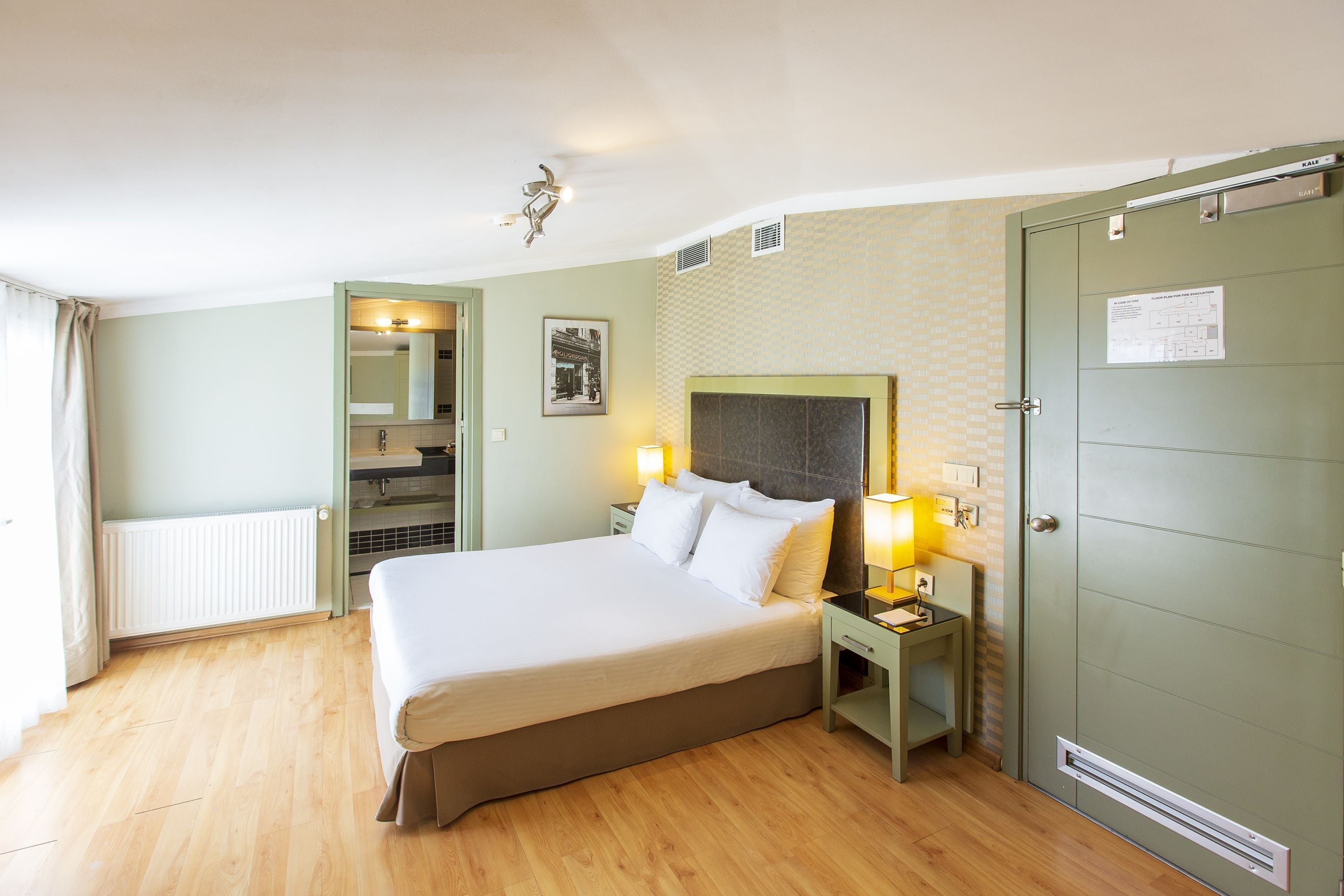 A hotel room with a bed, bedside lamps, a mirror, a radiator, and a door with a sign.
