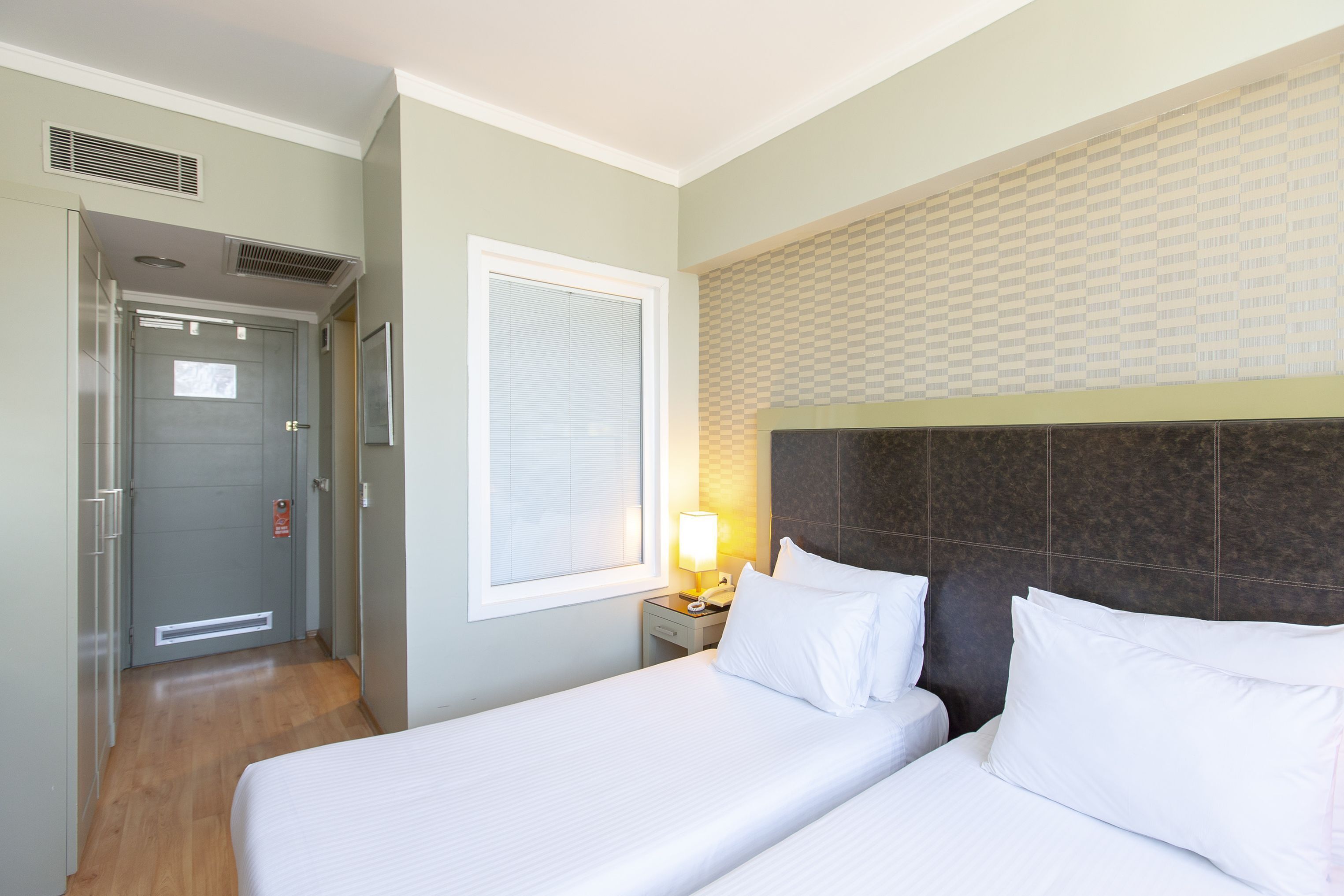 Een hotelkamer met twee bedden, een nachtkastje met een lamp, een raam met jaloezieën en een deur met ventilatie rooster.