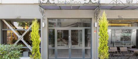 Entrada de hotel con puerta de vidrio, plantas en macetas y mesas al aire libre.