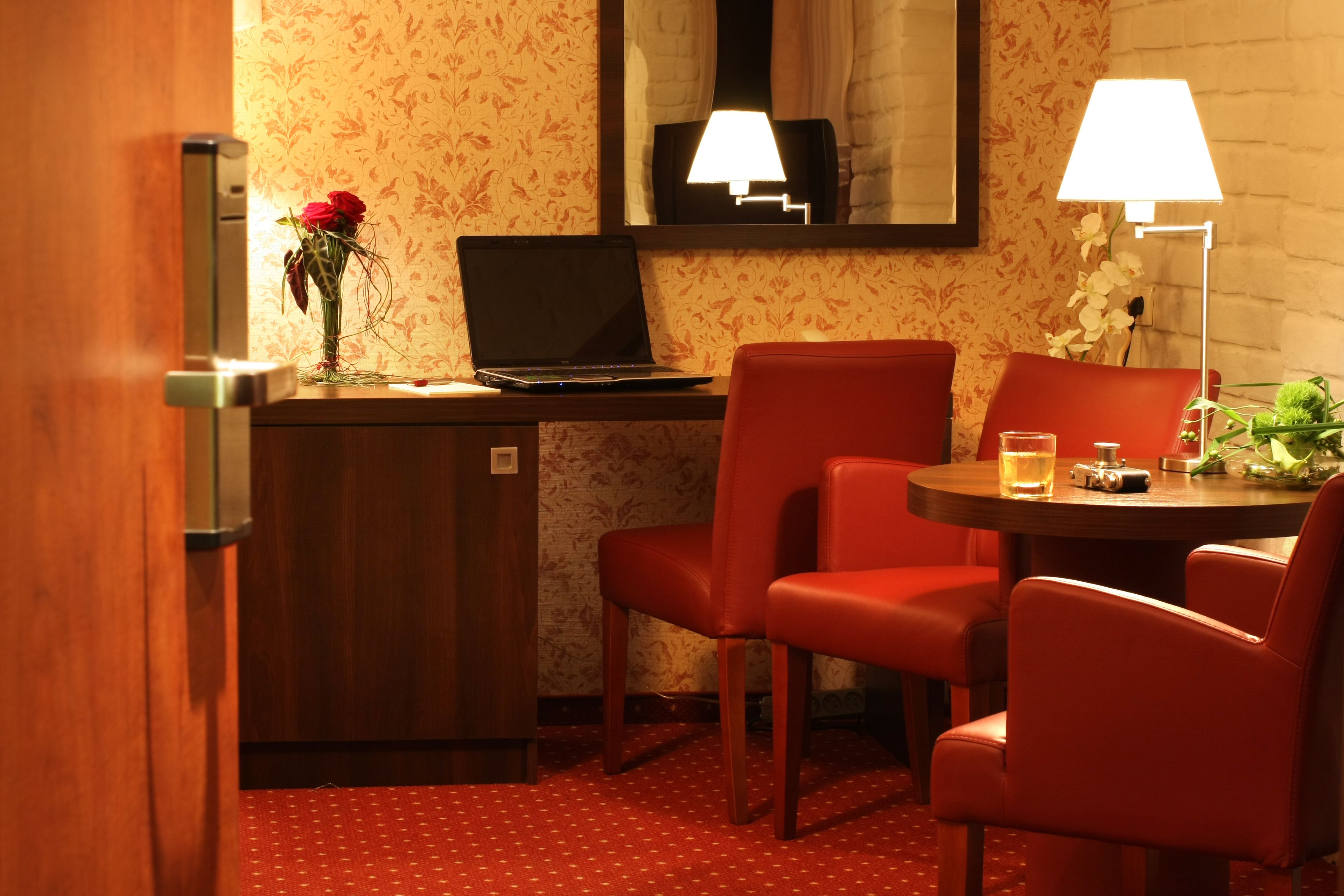 A hotel room with a desk, laptop, two red chairs, a small table with a glass and a camera, and a vase with flowers.