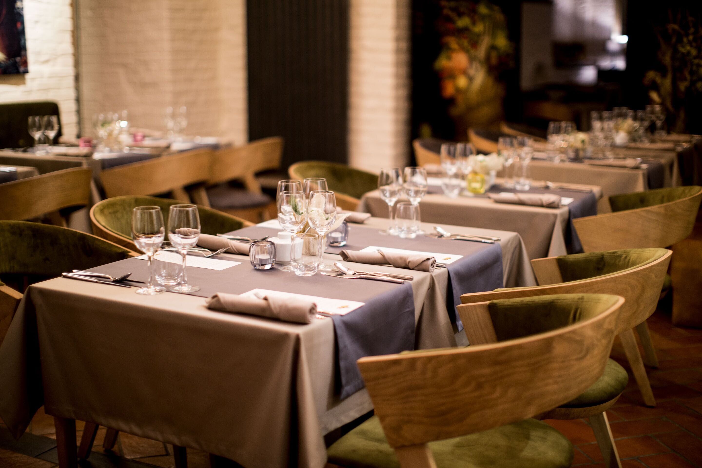 Un restaurant avec de nombreuses tables, chacune agrémentée de verres à vin et de couverts, de chaises en bois et d’un mur de briques en arrière-plan.