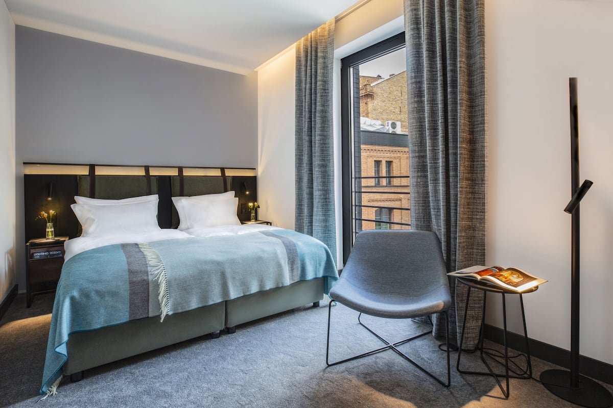 A hotel room with a bed, a chair, a nightstand, a floor lamp, and a window with curtains.