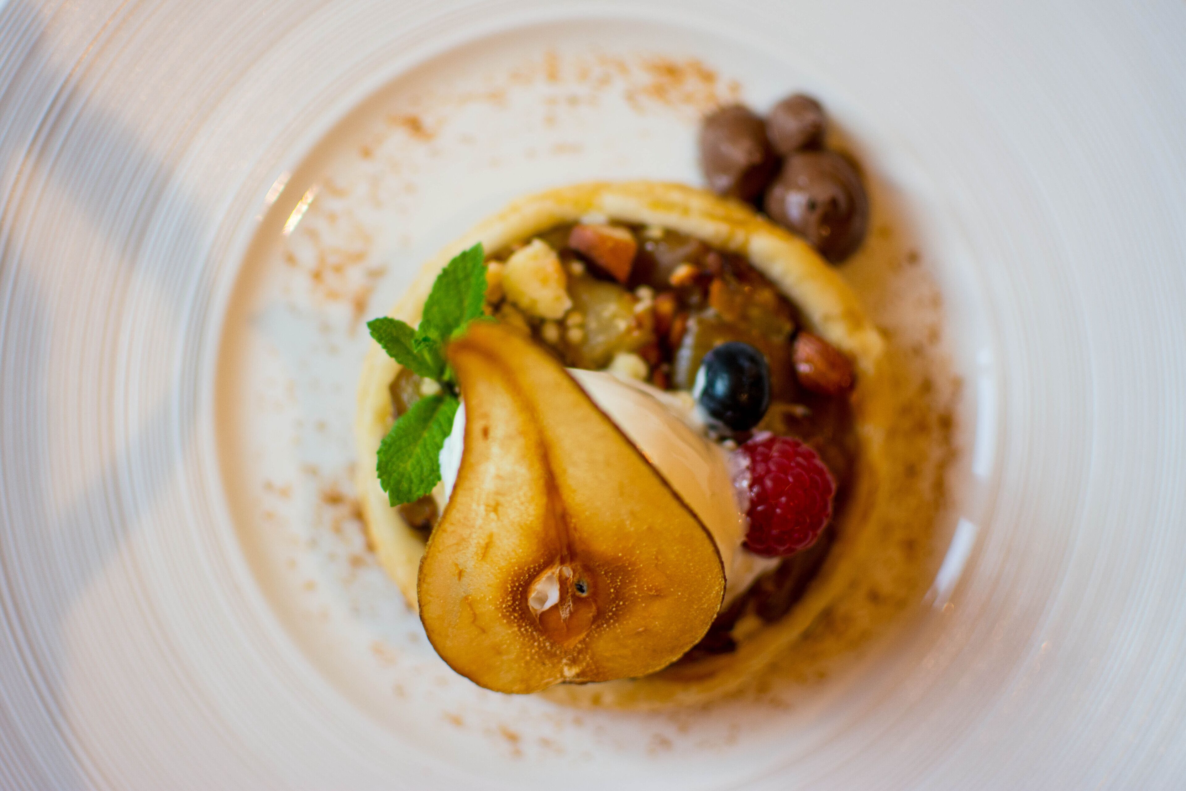 Un dessert con una pera, mirtilli, lamponi e un ciuffo di menta su un piatto bianco.