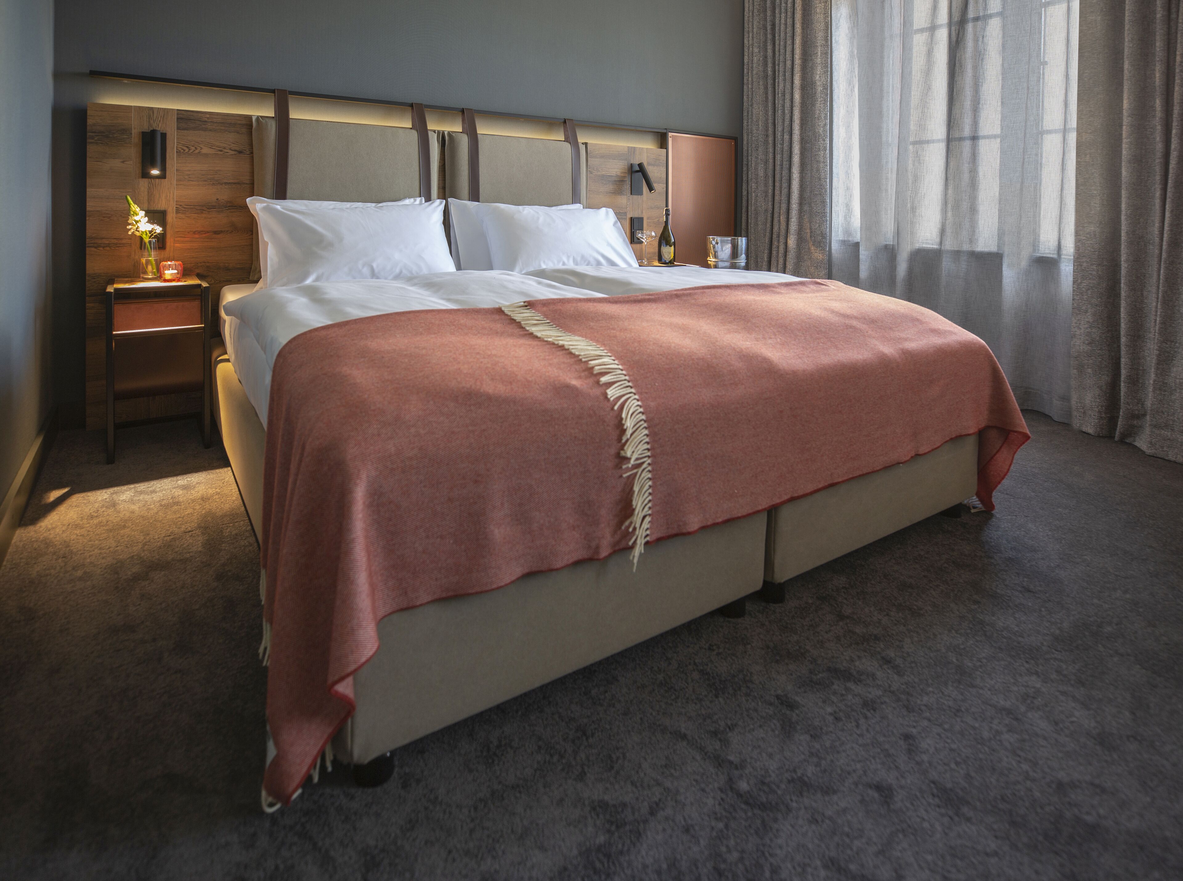Una cama bien hecha con un cabecero color beige, una manta roja y una mesita de noche con una lámpara y un florero.