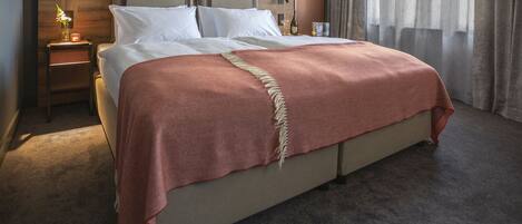 Una cama bien hecha con un cabecero color beige, una manta roja y una mesita de noche con una lámpara y un florero.