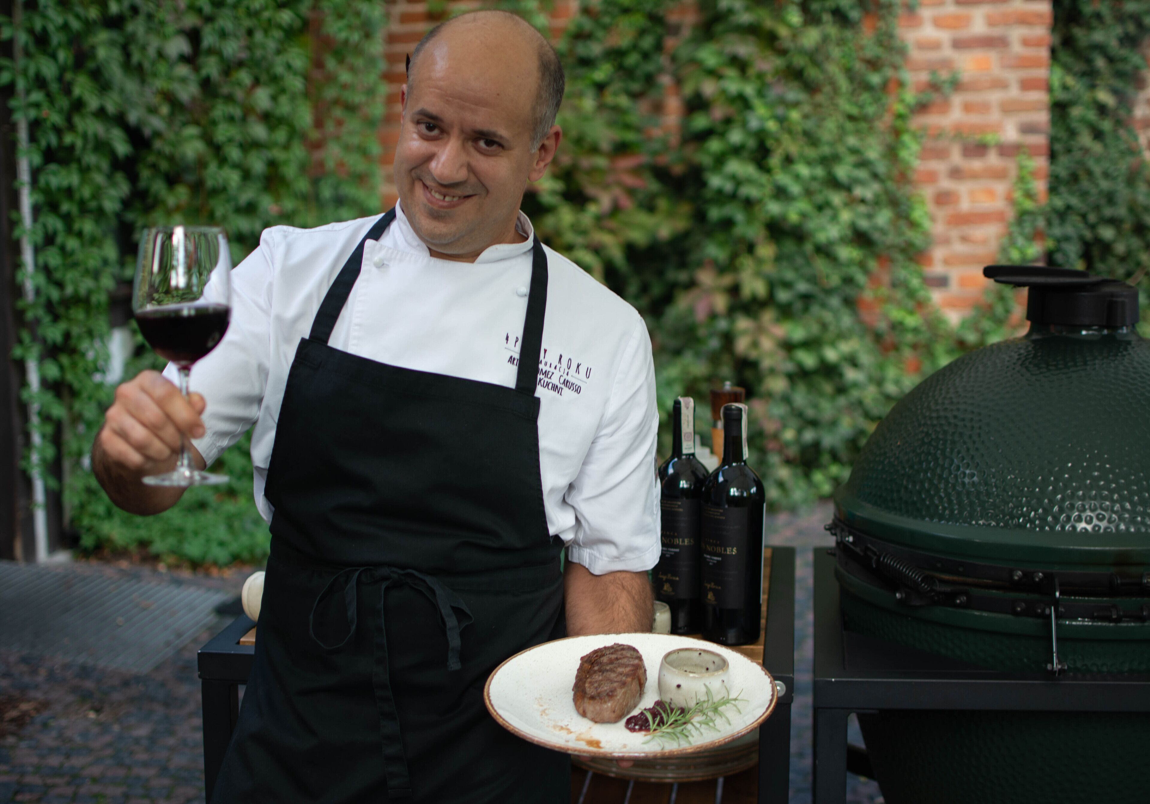 Un chef, tenant un verre de vin et une assiette de nourriture, se tient à côté d’un gril et de bouteilles de vin.