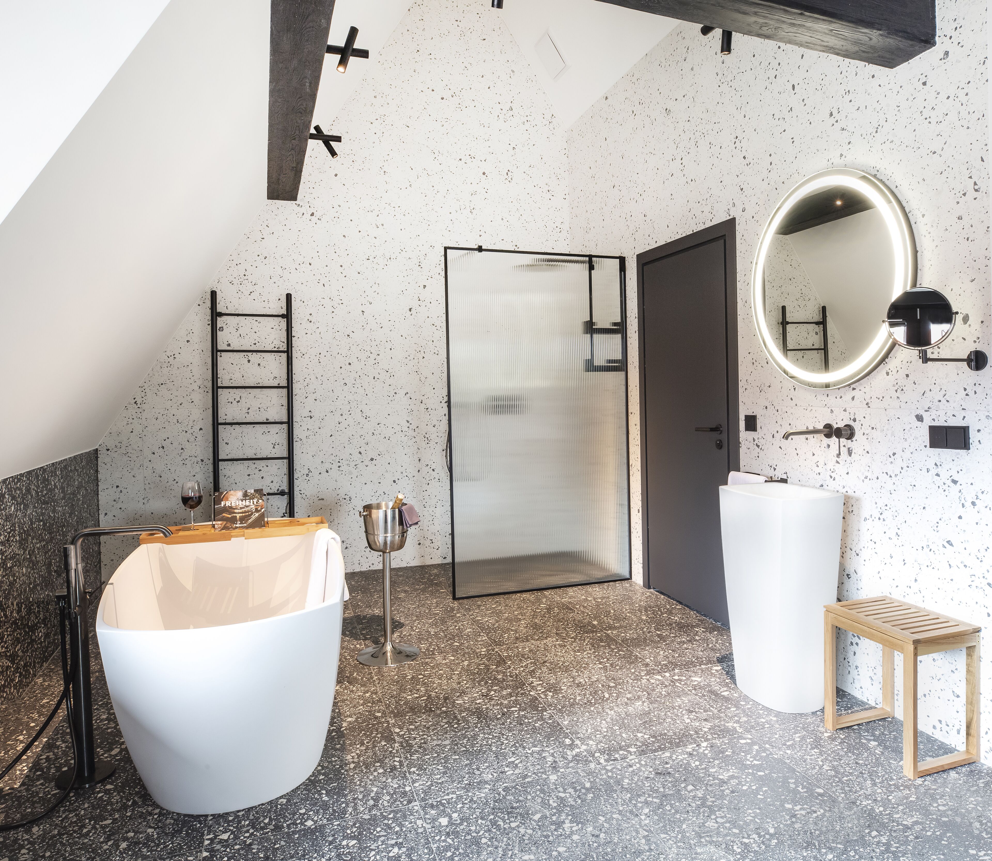 A modern bathroom with a freestanding bathtub, a shower enclosure, a round mirror, and a small wooden stool.