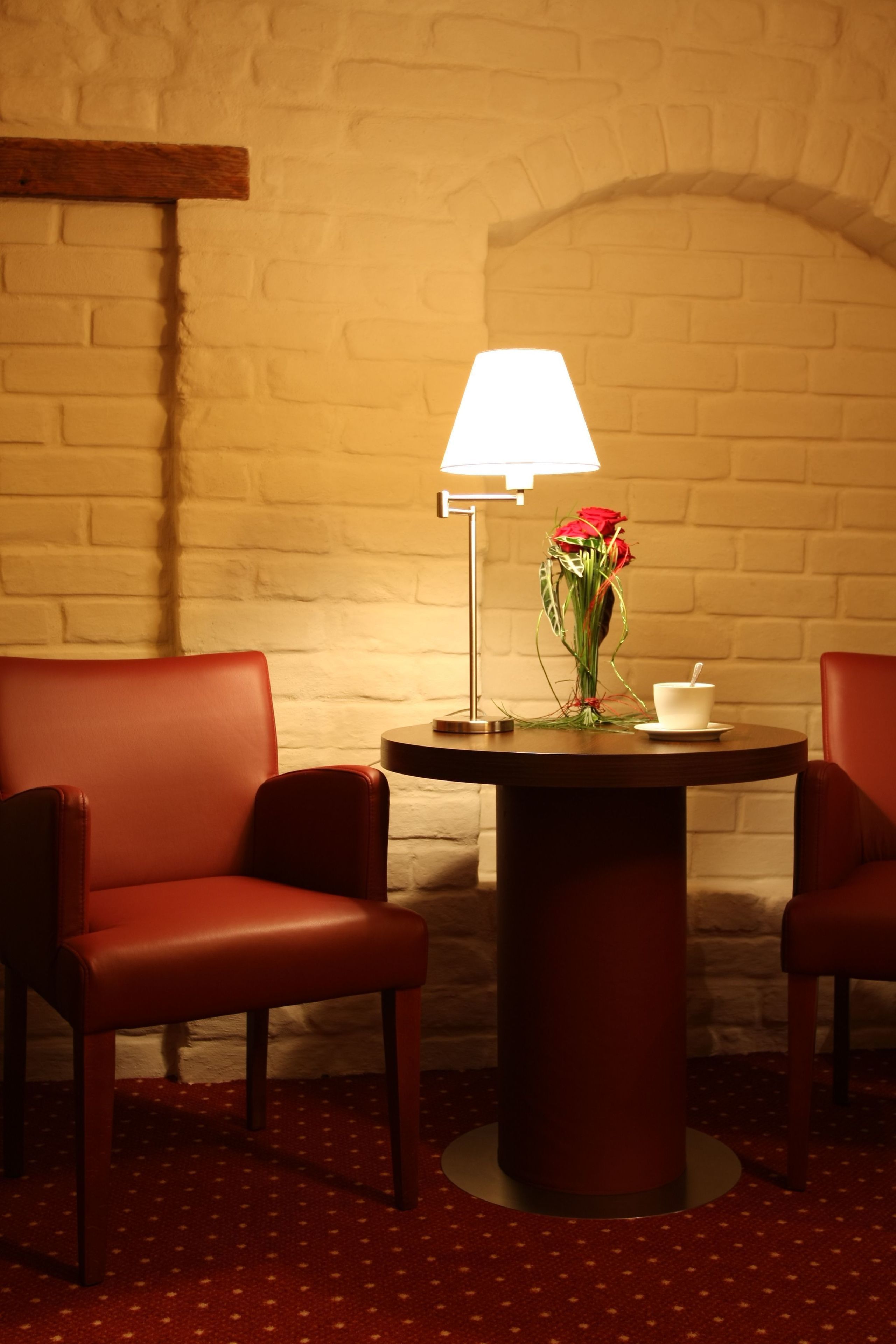 Una habitación con pared de ladrillo, dos sillas de cuero rojo, una mesa redonda con una lámpara de mesa blanca, un florero con flores rojas y una taza y platillo blancos.