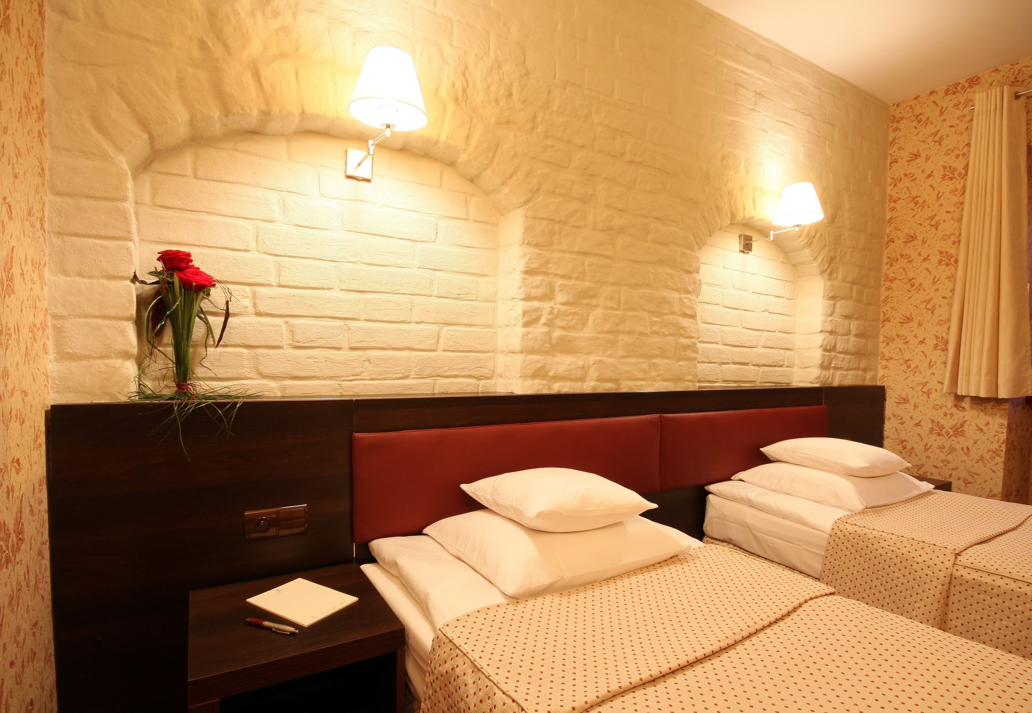 Habitación de hotel con pared de ladrillo, dos camas con ropa blanca, una mesita de noche con un bloc de notas y un florero con una flor roja.