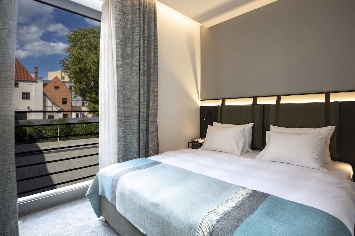 A hotel room with a large bed, a headboard with built-in lighting, a nightstand, and a view of buildings through a window with curtains.