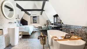 Un dormitorio moderno y bien iluminado con techo inclinado, una cama grande, un rincón de descanso con mesa y sillas, una bañera y un pequeño balcón con plantas.