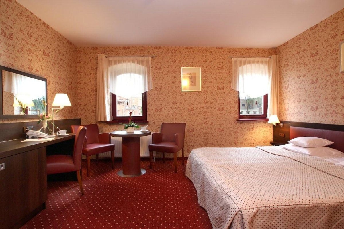 A hotel room with a bed, a desk with a telephone and a cup, two chairs, a table with a flower vase, and two windows with curtains.