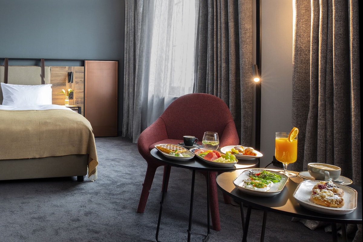 A hotel room with a large bed, a red armchair, and a breakfast table with plates of food and drinks.