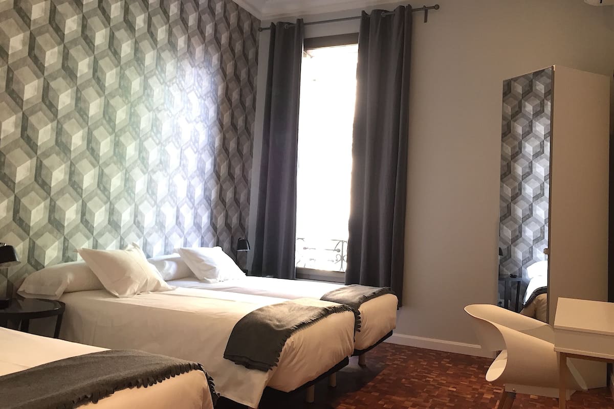 A hotel room with two beds, a desk, and a large window with curtains.