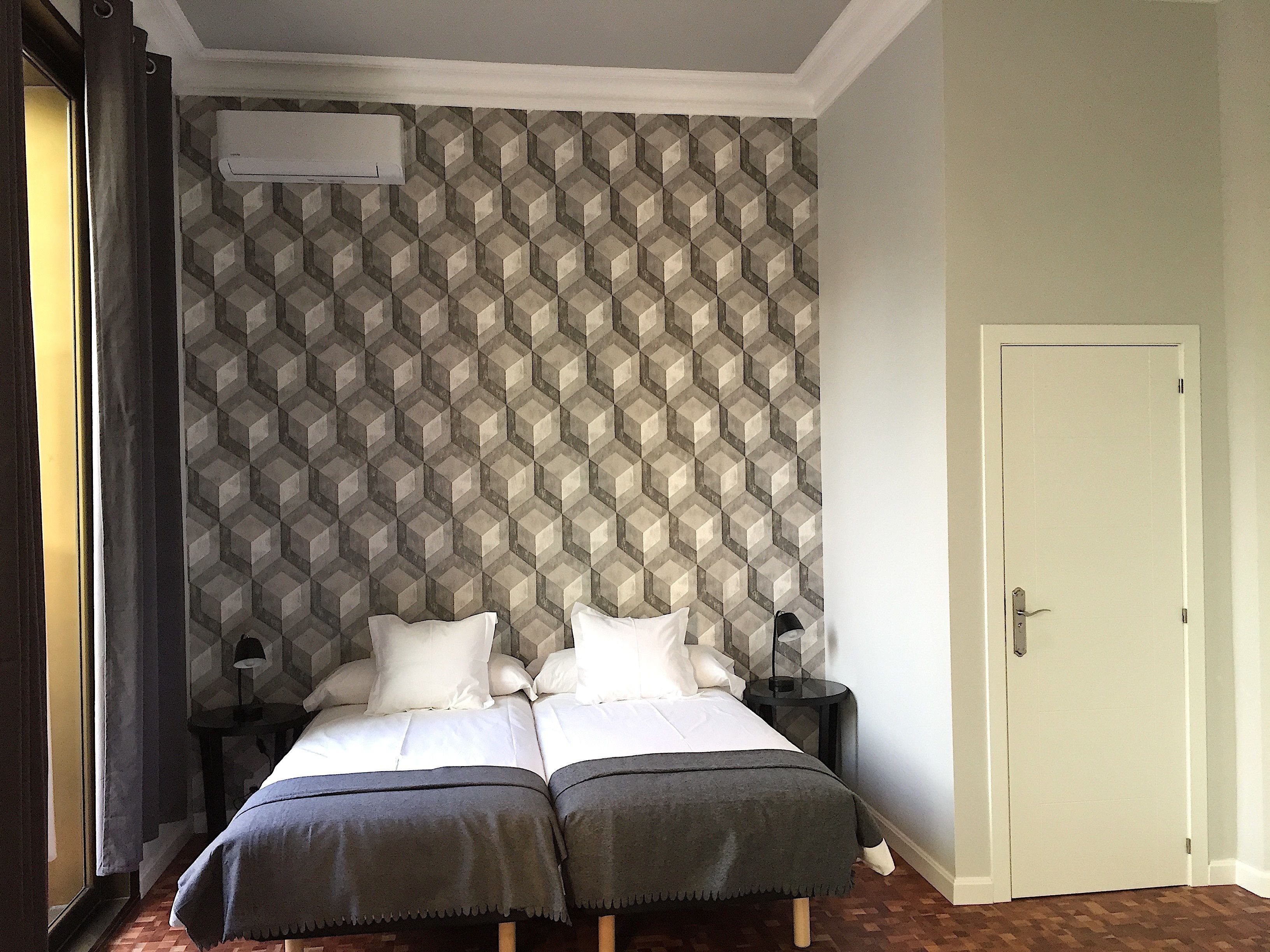 Una cama doble con dos almohadas y una colcha, una mesita de noche con una lámpara, un papel tapiz estampado y una puerta.