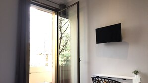 A bedroom with a bed, a desk, a chair, a TV, and a window with a view of trees and buildings. - Hip Karma Hostel (Barcelona)