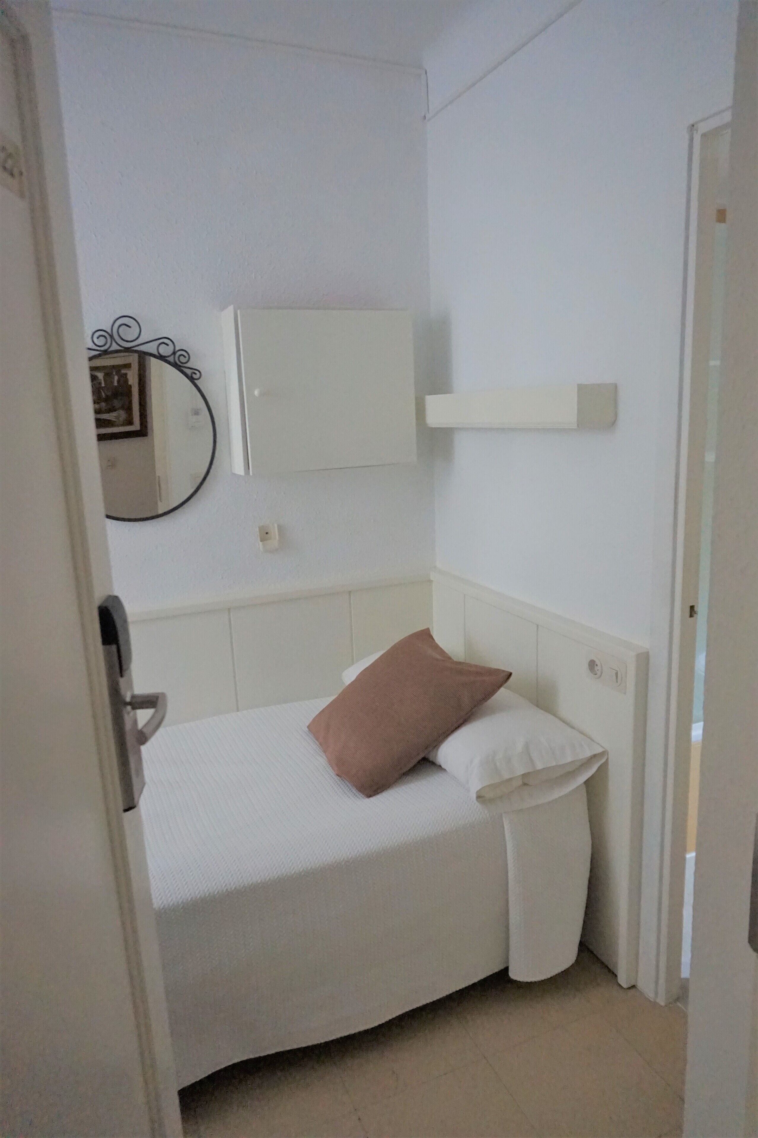 Un dormitorio pequeño y ordenado con una cama individual, una mesita de noche, un espejo y una estantería en la pared.
