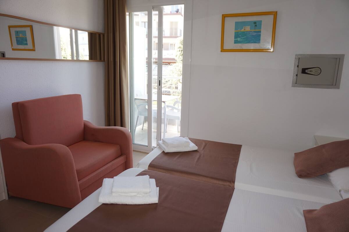 A hotel room with a red armchair, two beds with brown bedsheets, a white wall with a framed picture, and a window with a view of buildings.