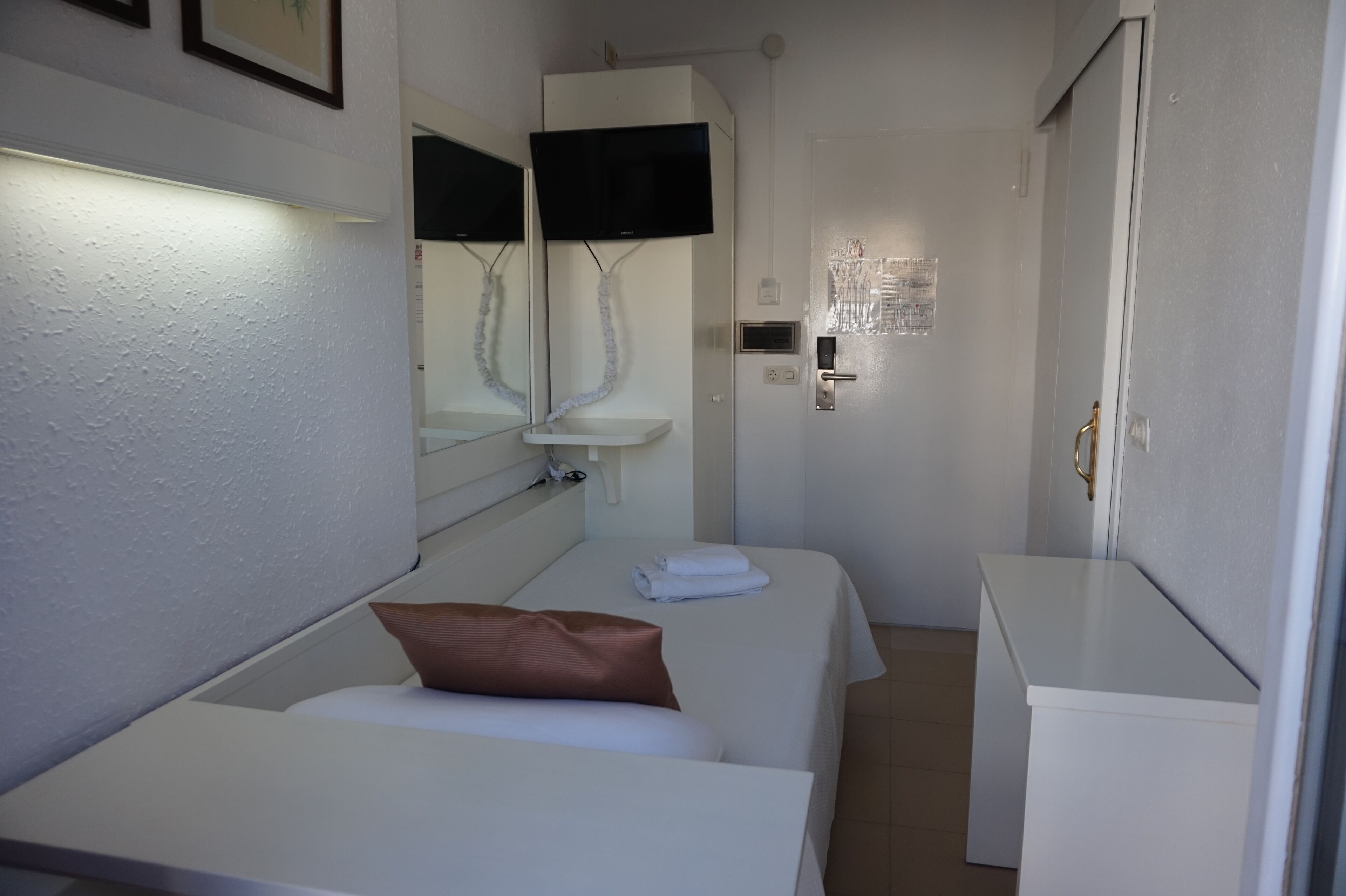 A compact hotel room with two single beds, a small white sink, a mirror, a television mounted above the sink, and a door with a handle.