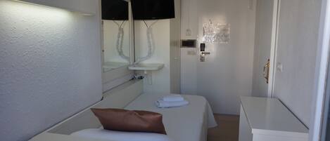 A compact hotel room with two single beds, a small white sink, a mirror, a television mounted above the sink, and a door with a handle.