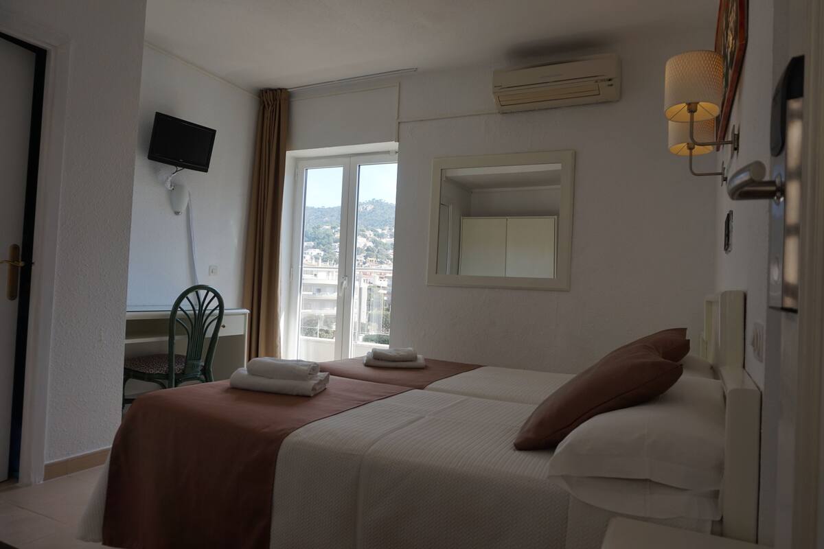 A hotel room with a bed, a desk, a chair, a TV, and a window with a view of buildings.