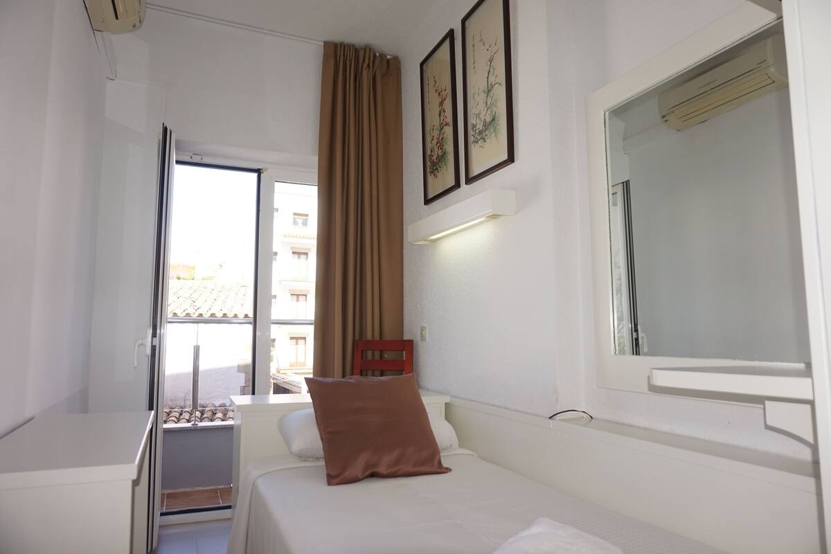 A hotel room with a bed, a window with curtains, a bedside table, and a wall-mounted air conditioning unit.
