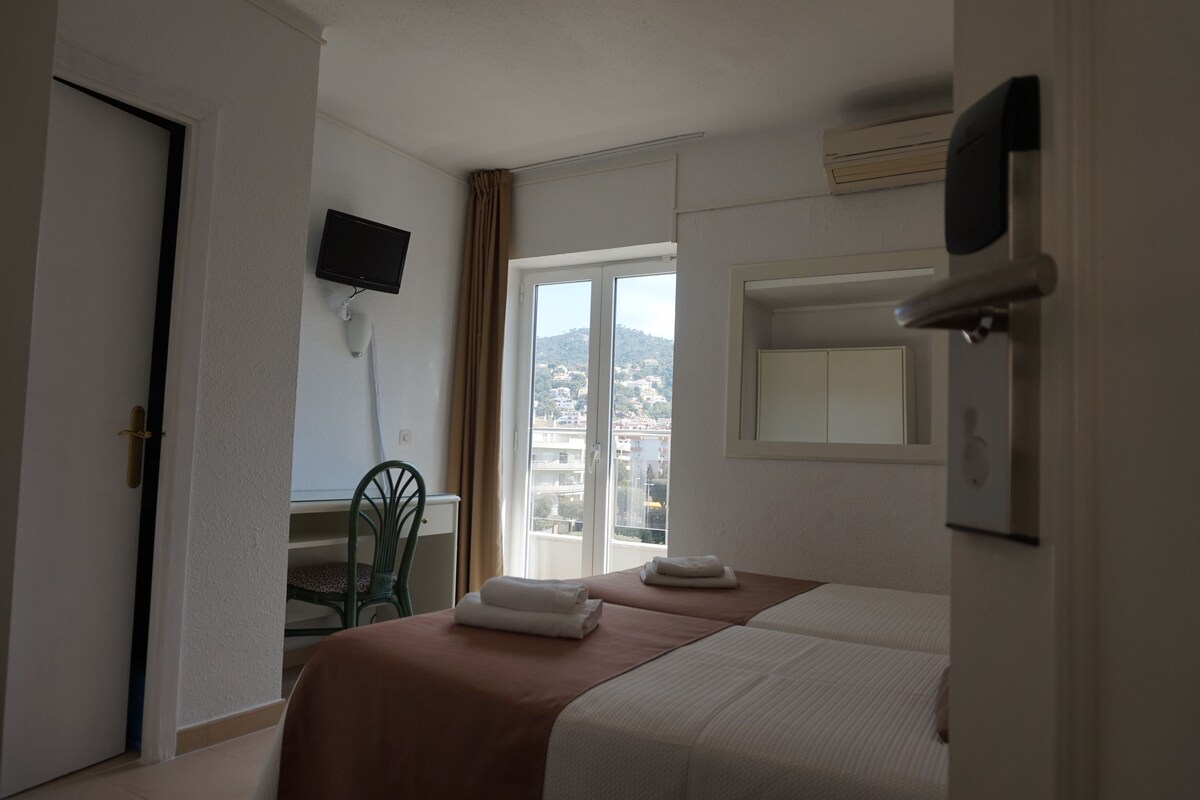 A hotel room with two beds, a TV mounted on the wall, a desk with a chair, and a view of the city through the window.