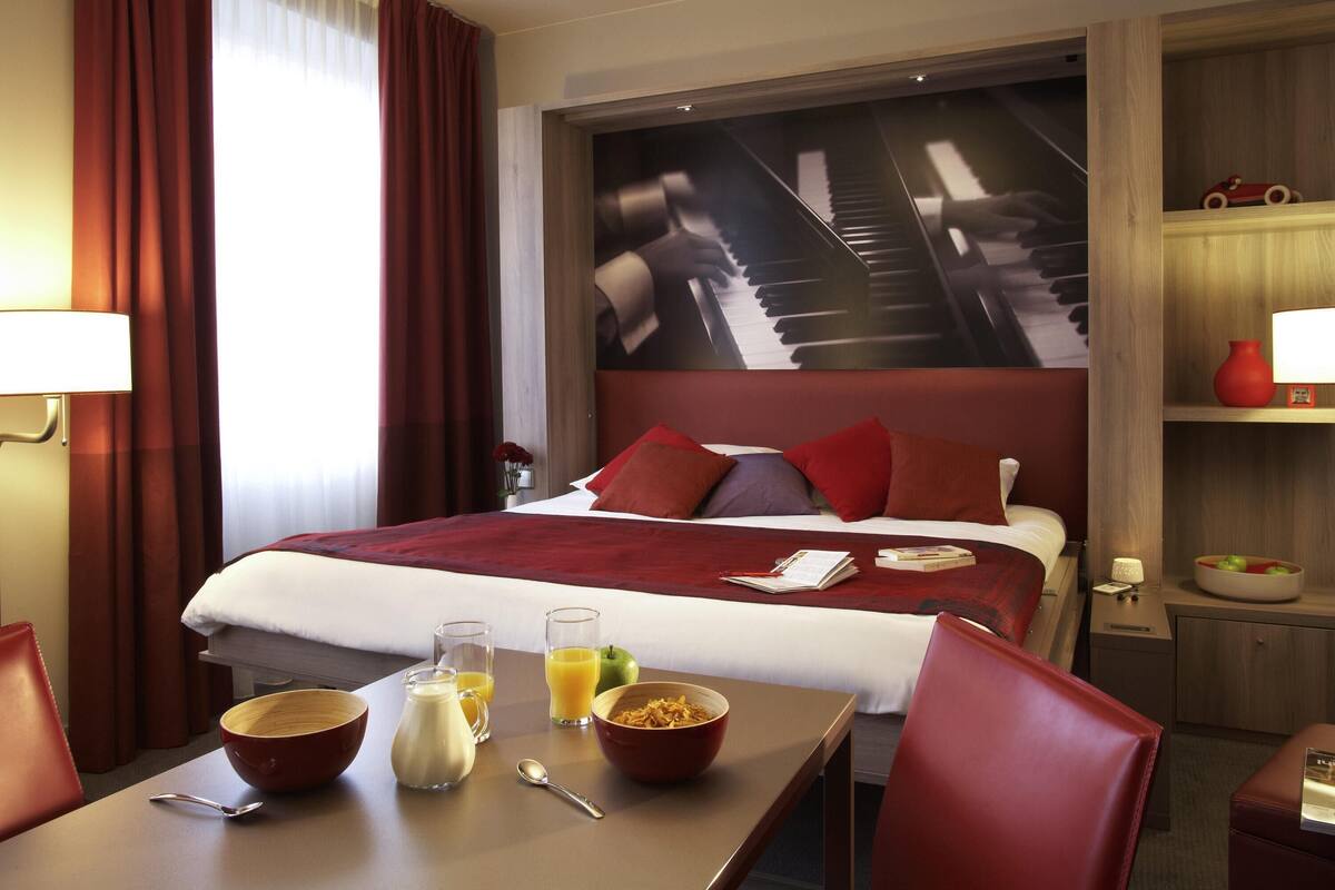 Una habitación de hotel con una cama, una mesa de desayuno con alimentos para el desayuno, una imagen de un piano en la pared y un estante pequeño con un jarrón.