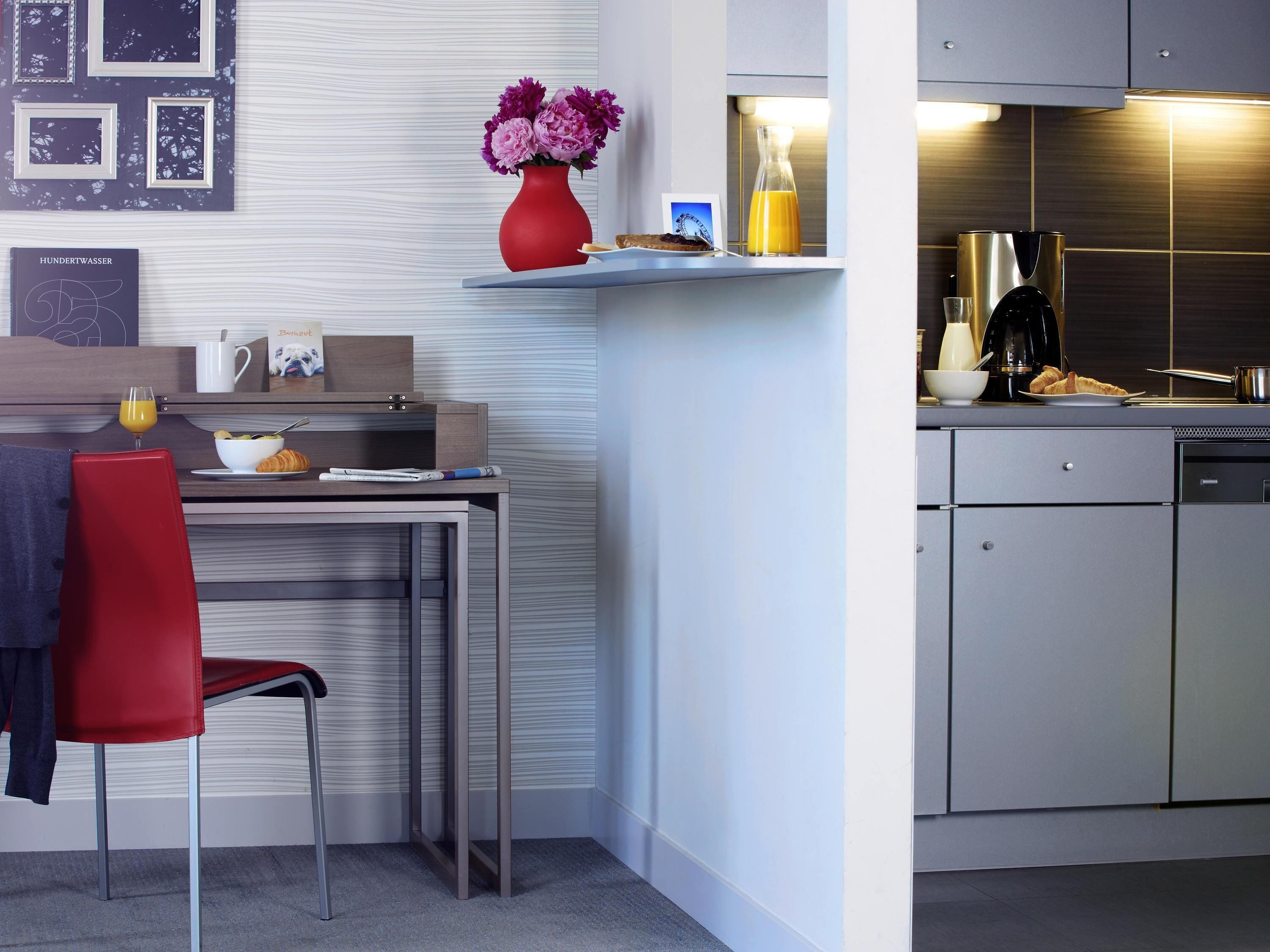 A modern kitchen with a built-in oven, a coffee machine, and a sink. There is a dining area with a red chair and a small table.