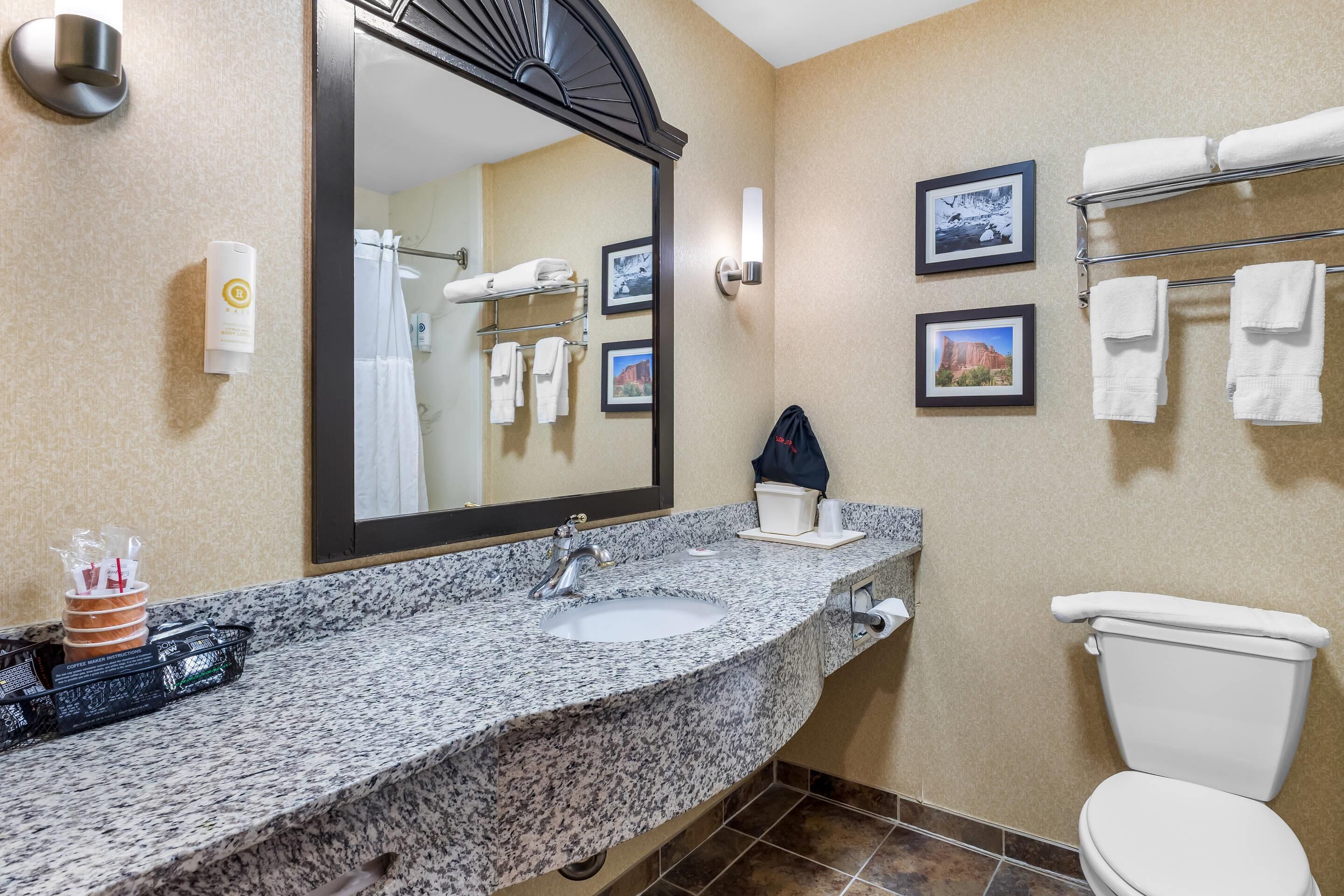 A bathroom with a granite countertop sink, a large mirror, a towel rack with towels, and a toilet.
