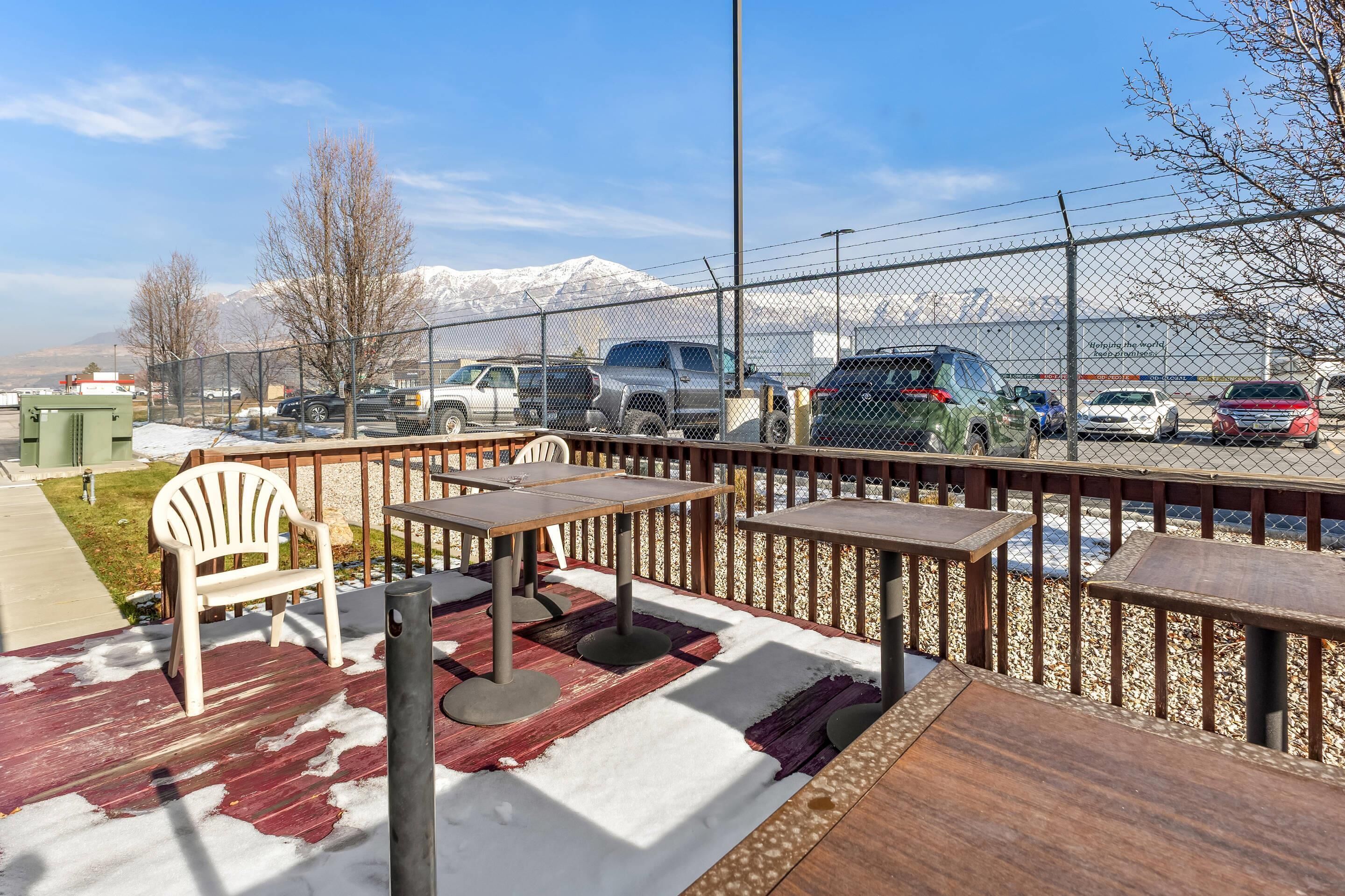 An outdoor seating area with tables and chairs, a wooden deck, and a view of a parking lot with cars and a mountain in the background.