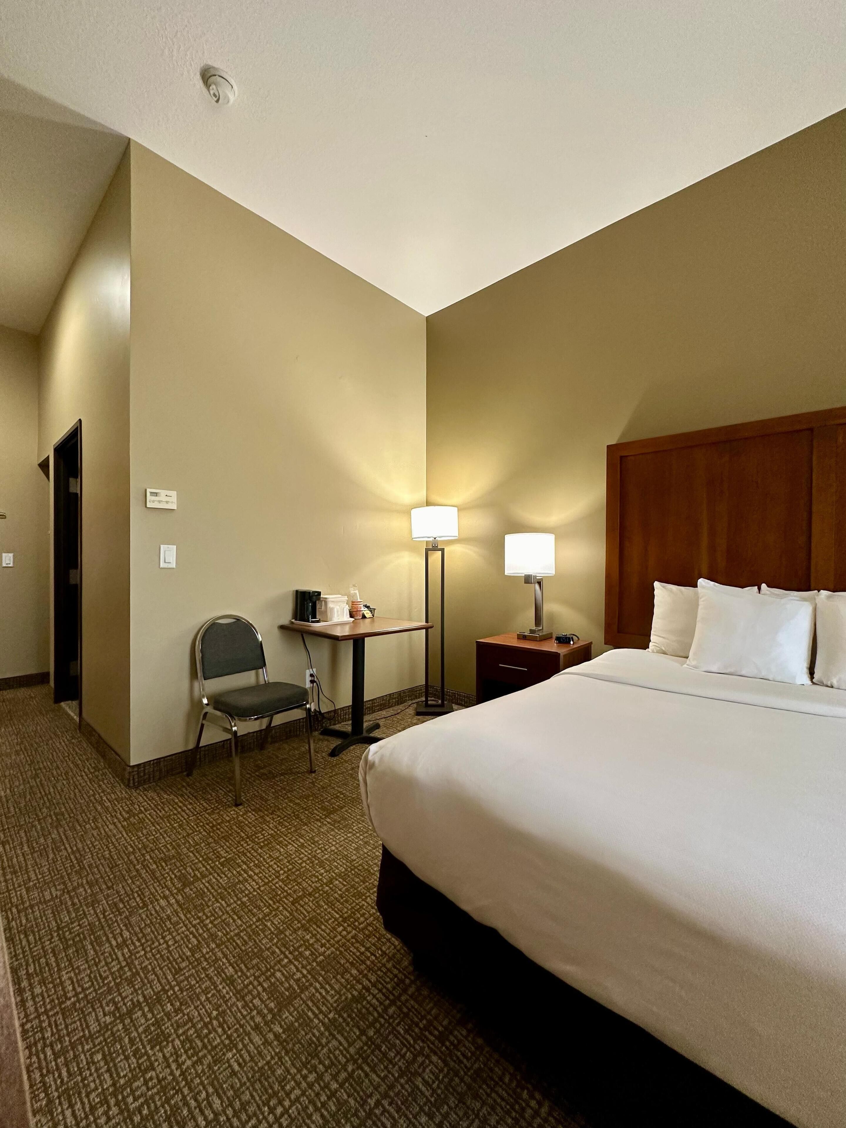 Una habitación de hotel con una cama grande, un escritorio con una silla, una lámpara y una pequeña mesa con una cafetera.