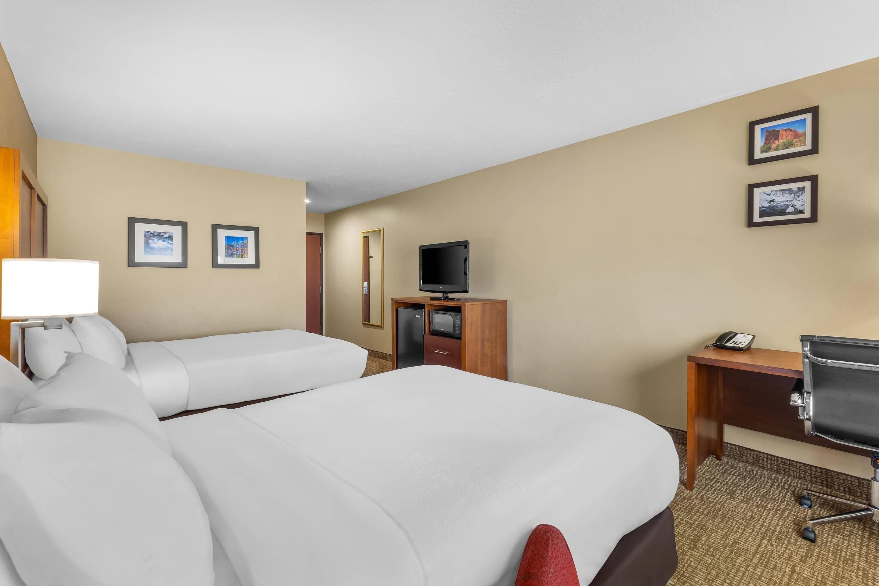 A hotel room with two beds, a desk, a chair, a TV, and a lamp.
