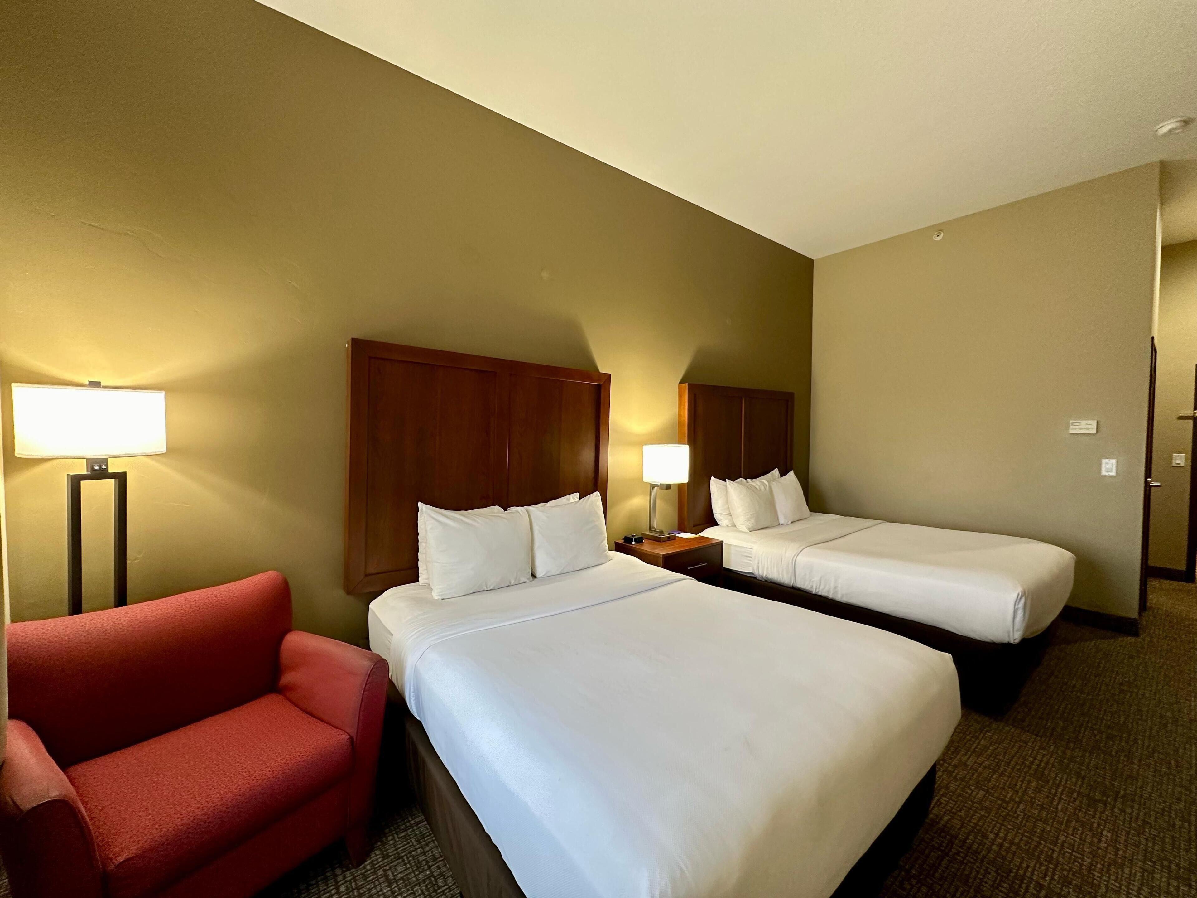 A hotel room with two beds, a red armchair, and a nightstand with a lamp.