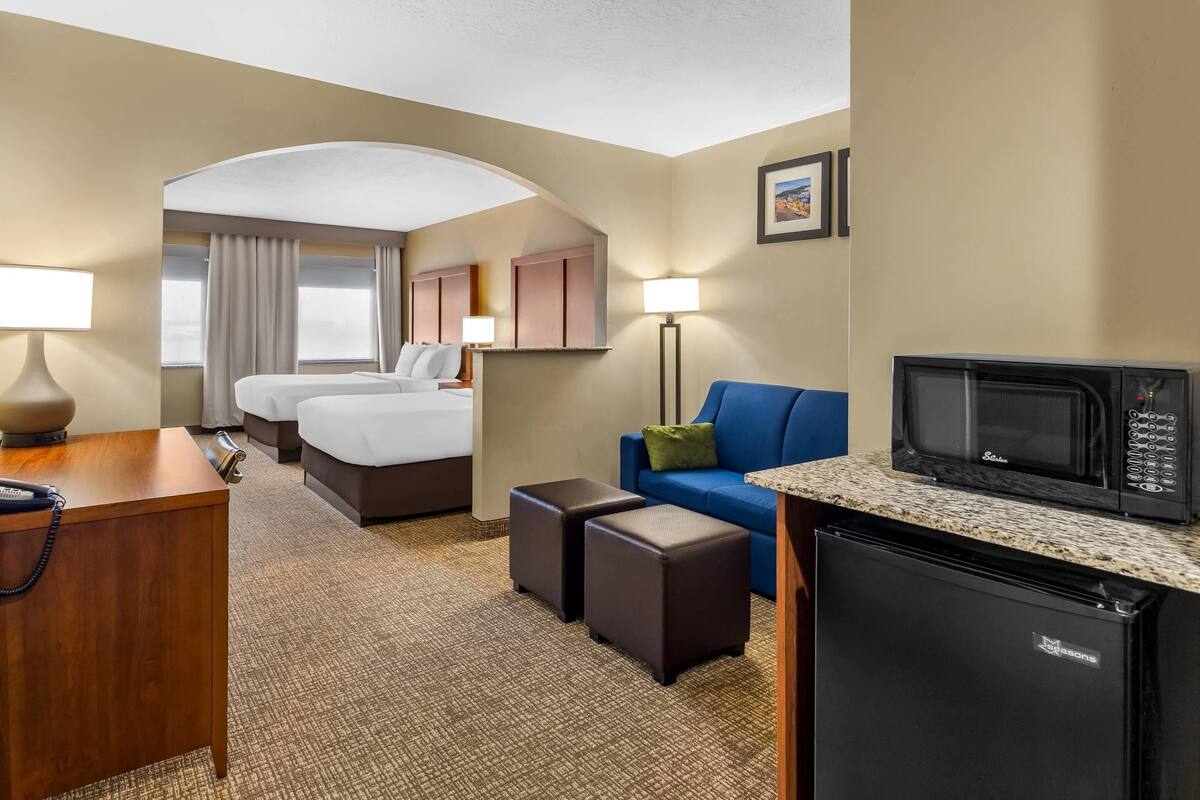 A hotel room with a bed, two bedside tables, a desk with a telephone, a microwave, a refrigerator, a sofa, and a small table with a lamp.