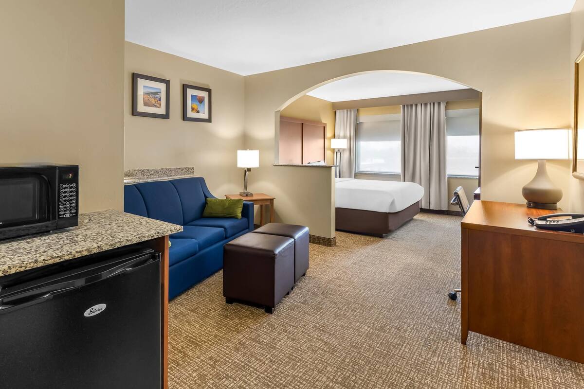 A hotel room with a kitchenette, a bed, a desk, a blue sofa, and a small table with a lamp.