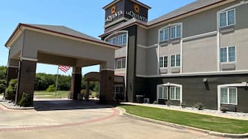 La Quinta's elegant entrance features a covered walkway and an American flag, set against a backdrop of large windows and landscaped gardens.