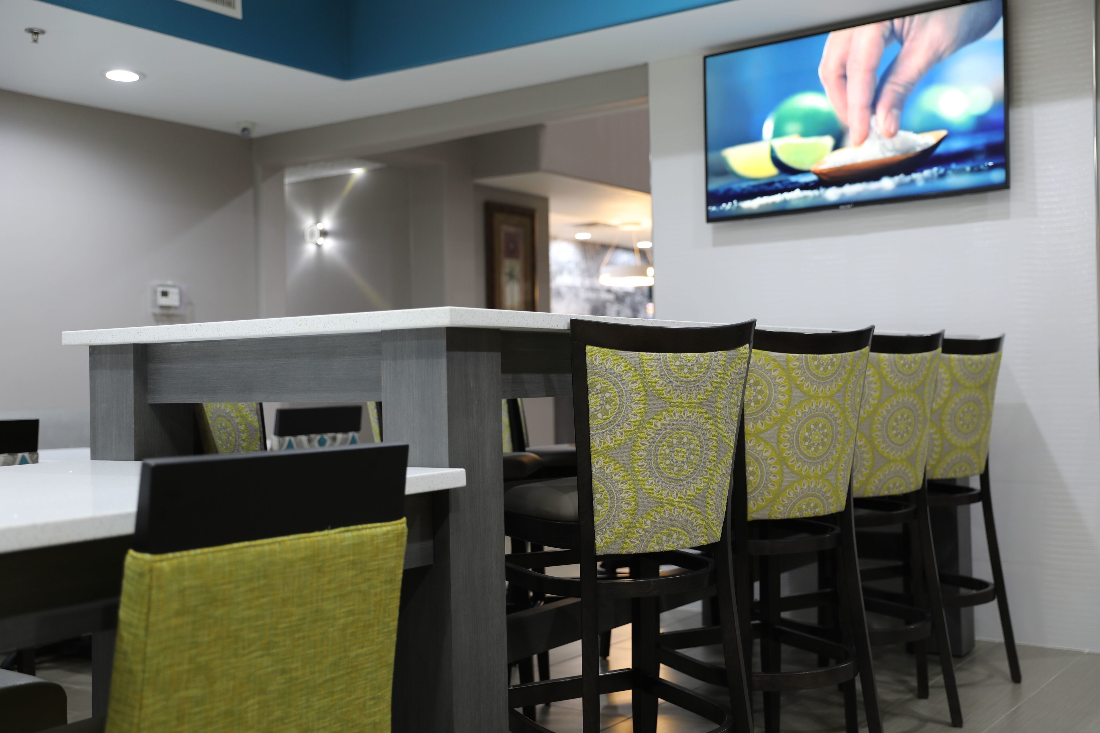 Un espace repas moderne avec une longue table et des chaises, un téléviseur à écran plat fixé au mur et une nappe à motifs.