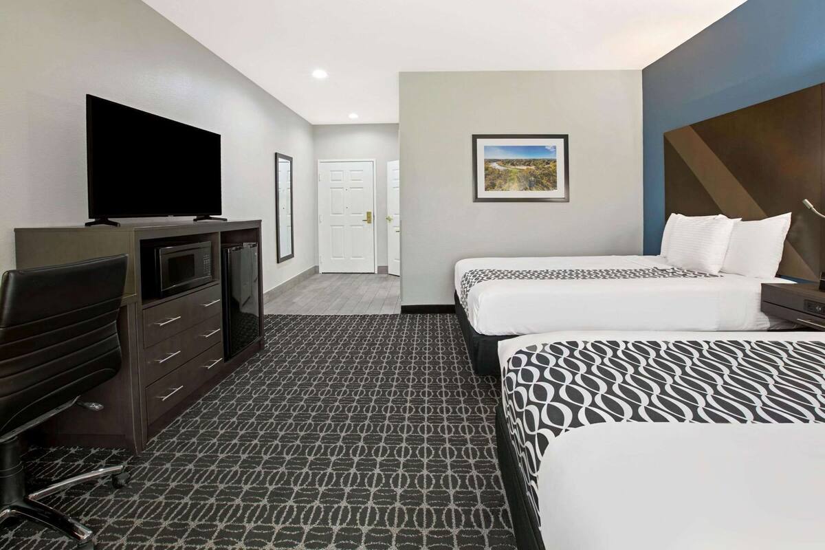 Une chambre d’hôtel comprenant un lit, un bureau avec une télévision, une commode, un tableau encadré au mur et une porte donnant sur une autre pièce.