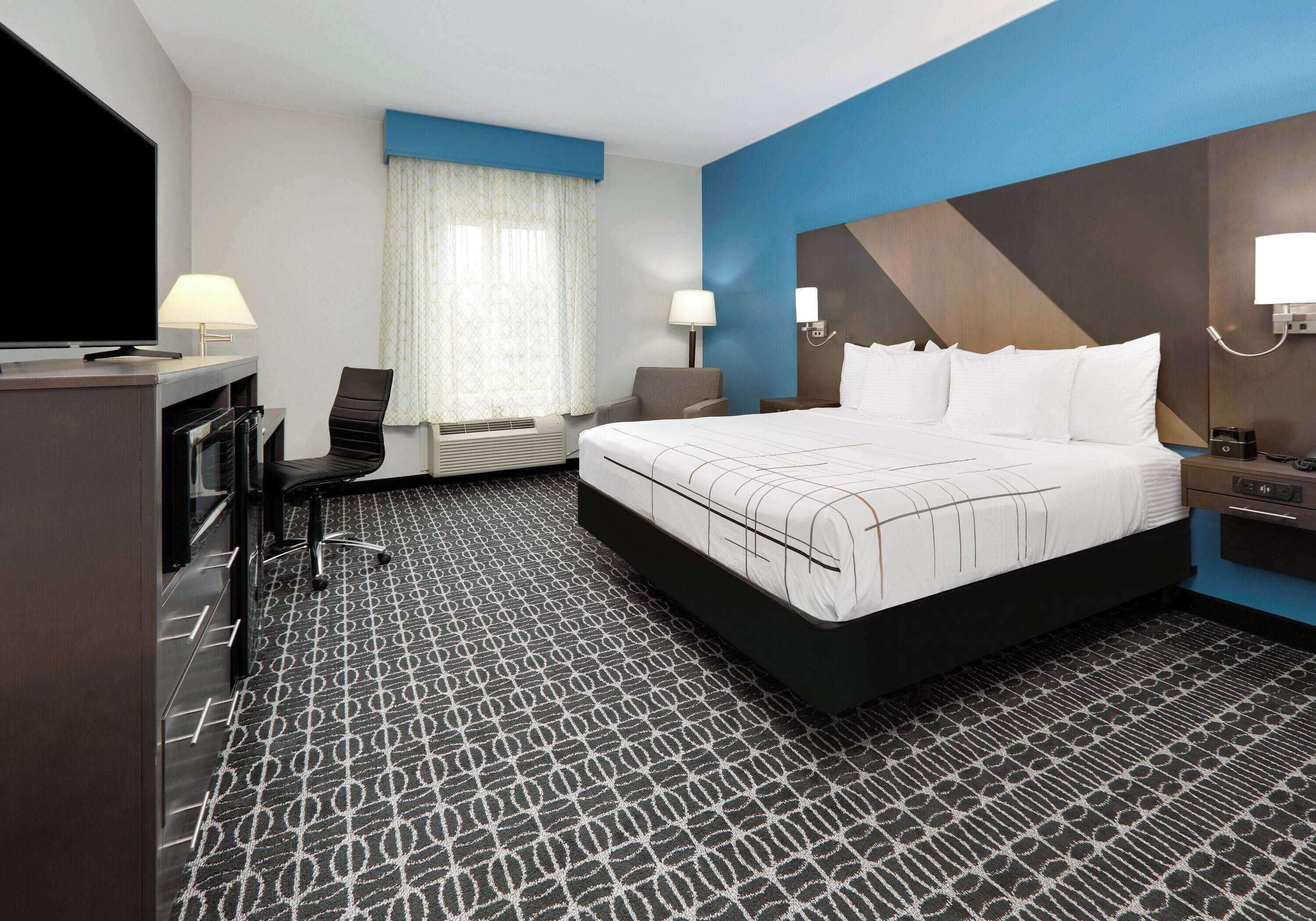 A hotel room with a large bed, a desk with a flat-screen TV, a chair, a desk lamp, and a window with curtains.