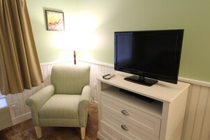 Quarto com uma poltrona verde, um móvel branco com TV de tela plana, uma luminária e um quadro na parede.