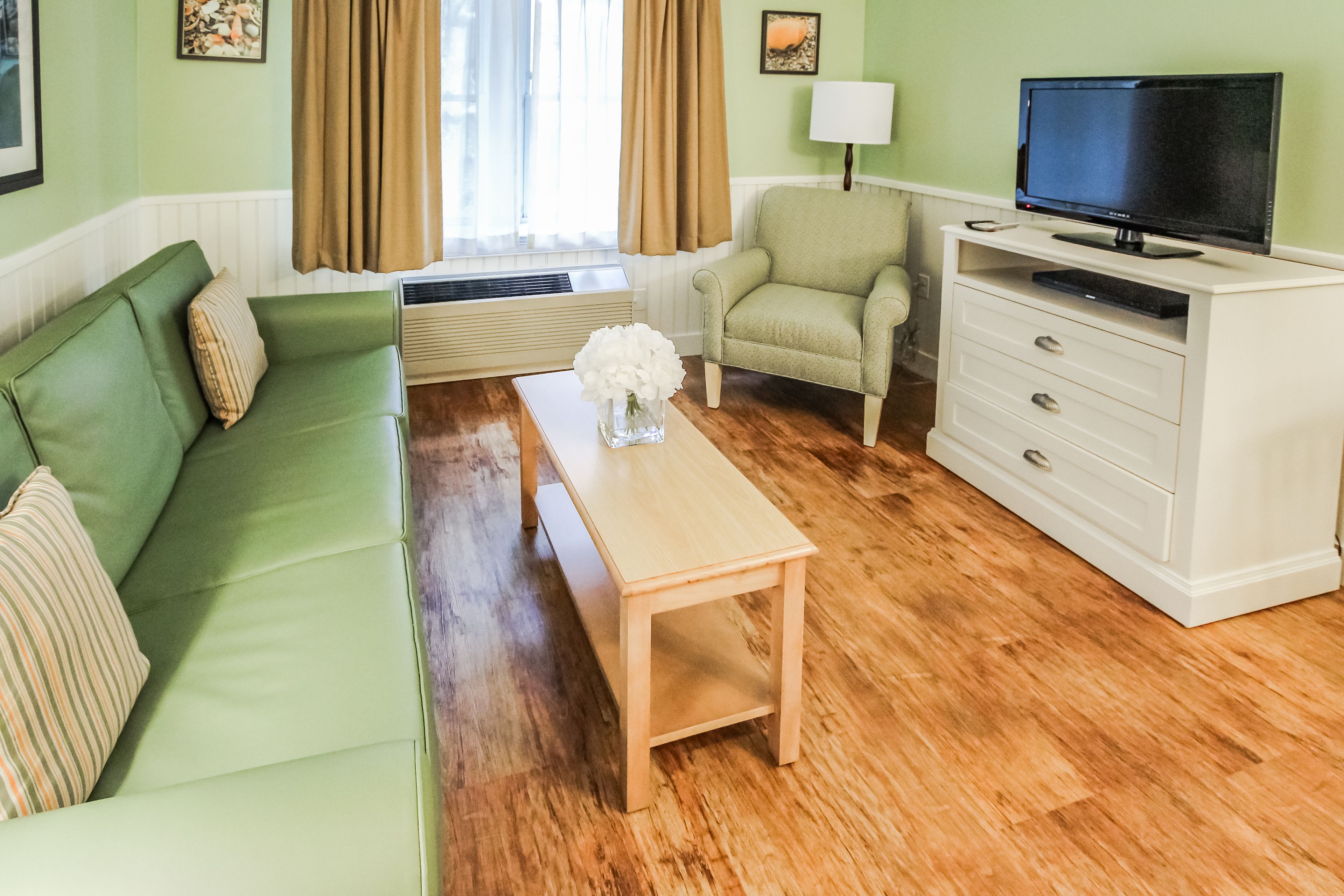 Un salon avec un canapé vert, une table basse en bois, un meuble blanc avec un téléviseur et un fauteuil beige.