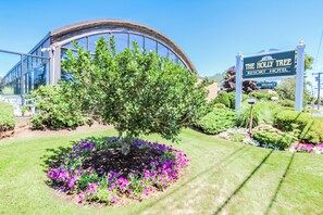 A modern building with a curved glass facade, a sign indicating "The Holly Tree Resort Hotel," and a well-maintained garden with purple flowers and a large tree.