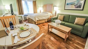 Habitación de hotel con cama, sofá verde, mesa de comedor preparada para dos personas y un cuadro en la pared.