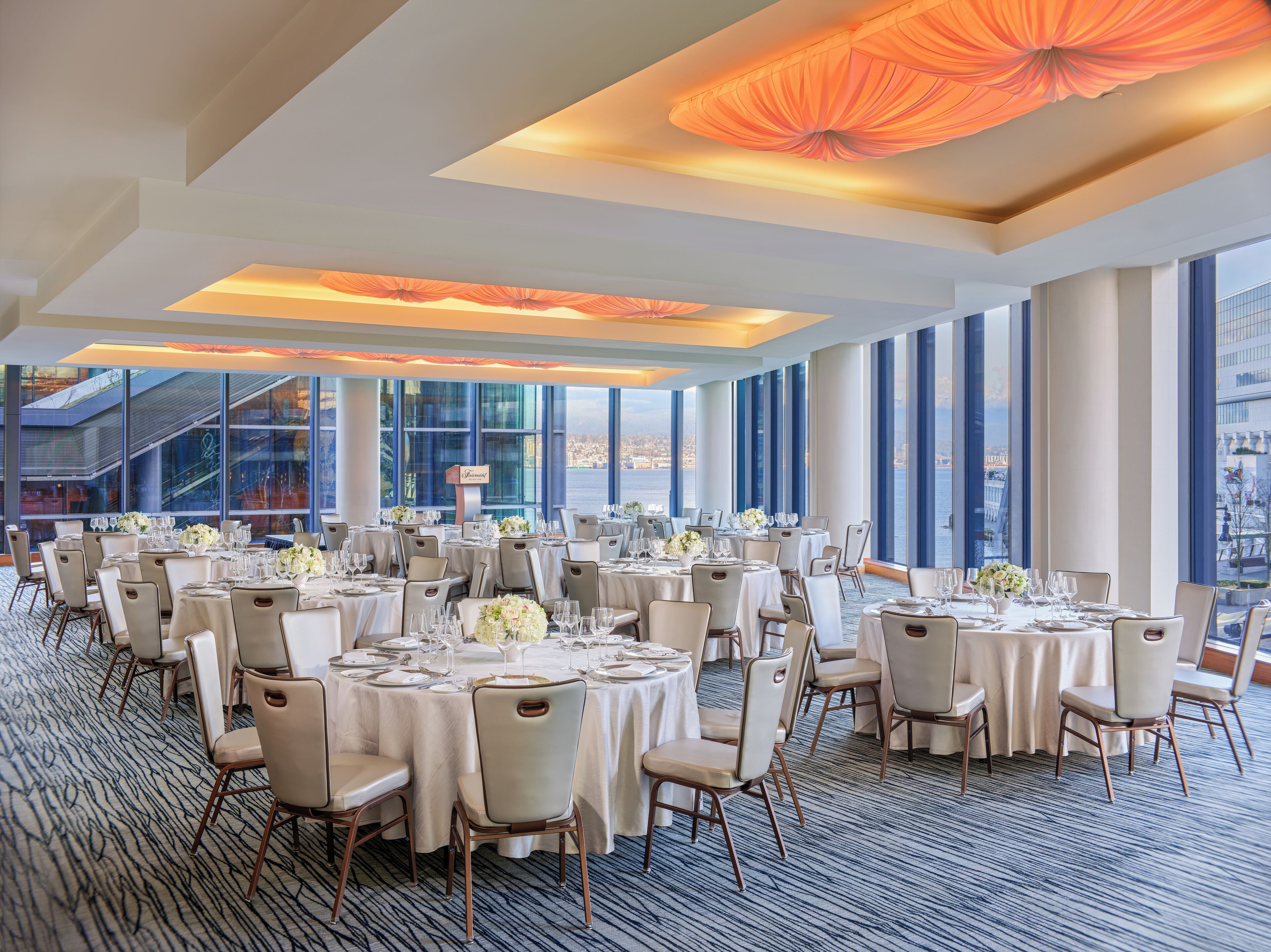 A spacious banquet hall with round tables set for dining, large windows offering a city view, and a modern ceiling design.