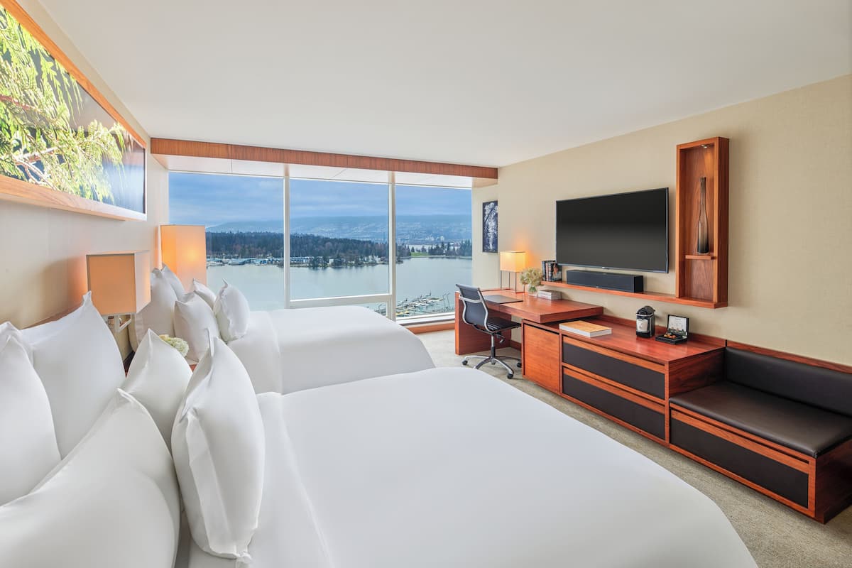 A hotel room with two beds, a desk, a TV, and a large window offering a view of a body of water and a cityscape.