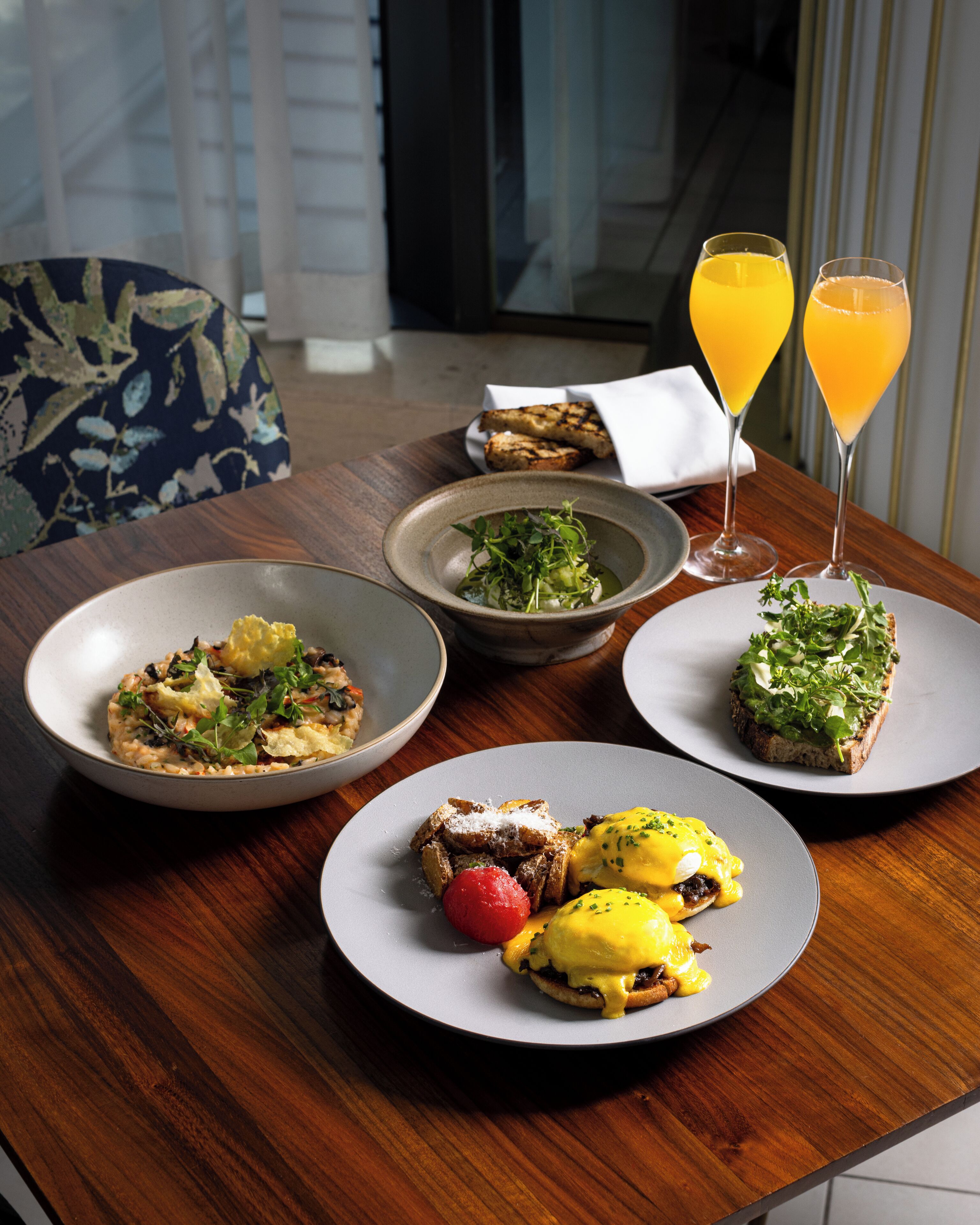 A dining table with various dishes, including eggs benedict and toast with greens, and two glasses of orange juice.